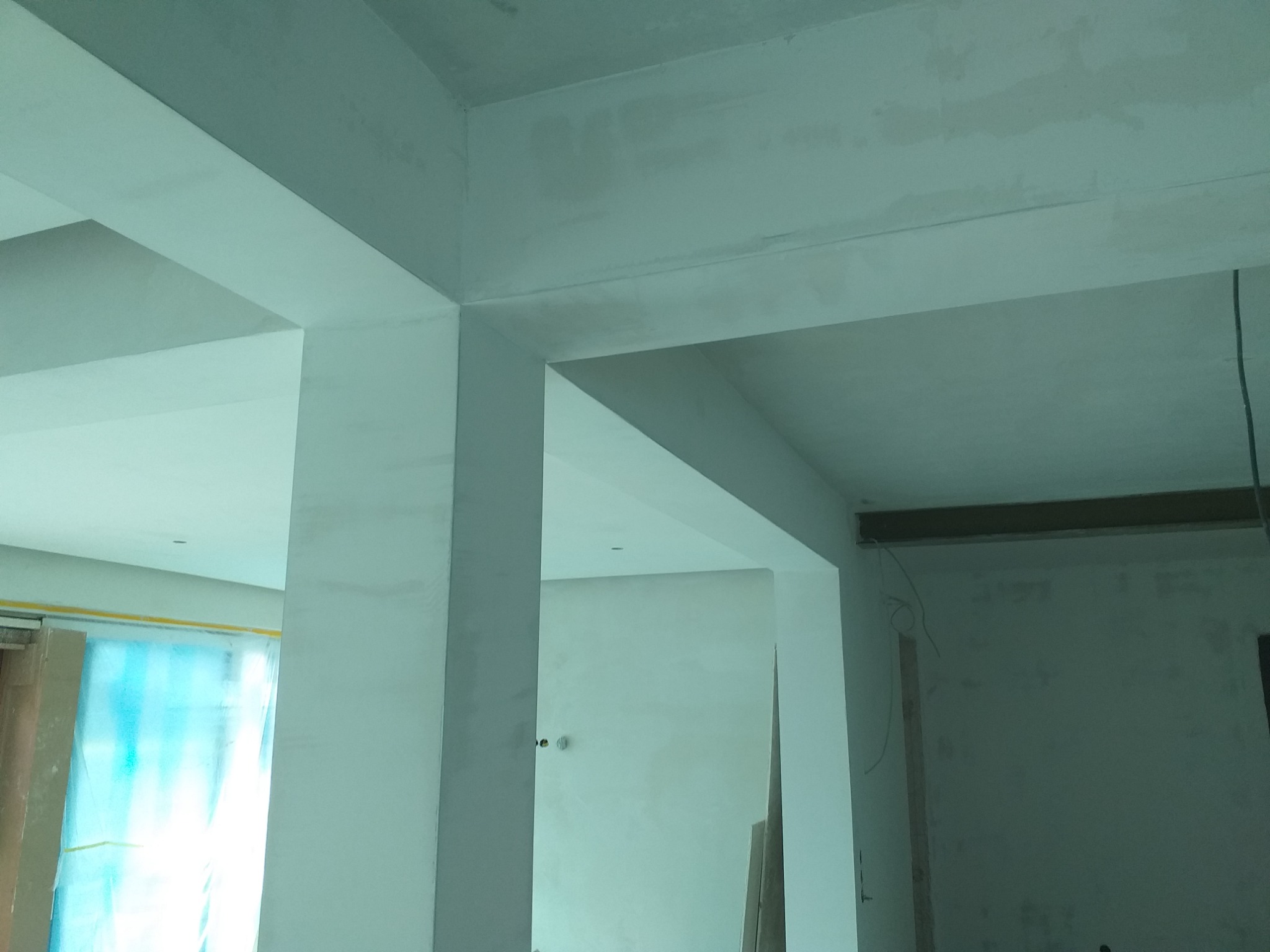 Wnętrze w trakcie malowania: sufit, ściany i filary pokryte świeżą, jasną farbą, widoczne niedoskonałości i ślady po szpachlowaniu, zabezpieczona folią stolarka okienna w tle.