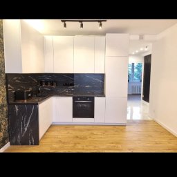 mak.bud.nowak - Nowoczesna kuchnia z białymi szafkami i blatem z czarnego marmuru, oświetlenie szynowe, drewniana podłoga, widok na dalszą część mieszkania.