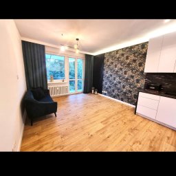 mak.bud.nowak - Wnętrze pokoju po remoncie: drewniana podłoga, ściana z tapetą w kwiatowy wzór, ciemnozielone zasłony i sofa, białe meble kuchenne z czarnym blatem.