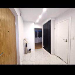 mak.bud.nowak - Wykończony korytarz w Opolu z białymi marmurowymi płytkami podłogowymi, drewnianymi drzwiami wejściowymi, białymi ścianami, czarnym panelem dekoracyjnym i białymi drzwiami wewnętrznymi.