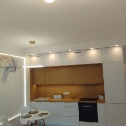 mak.bud.nowak - Nowoczesna kuchnia z białymi szafkami, drewnianymi frontami, okrągłym stołem i dekoracyjną tapetą imitującą marmur z niebiesko-złotymi żyłami, oświetlona lampą wiszącą i ledowym paskiem.