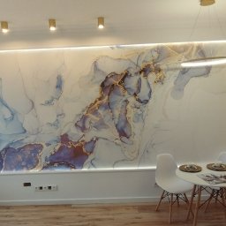 mak.bud.nowak - Nowoczesne wnętrze mieszkania z podświetlaną ścianą z abstrakcyjnym wzorem w odcieniach błękitu, złota i fioletu, mały okrągły stolik z dwoma krzesłami, fragment zabudowy kuchennej z drewnianymi...