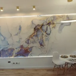 Nowoczesne wnętrze mieszkania z podświetlaną ścianą z abstrakcyjnym wzorem w odcieniach błękitu, złota i fioletu, mały okrągły stolik z dwoma krzesłami, fragment zabudowy kuchennej z drewnianymi...