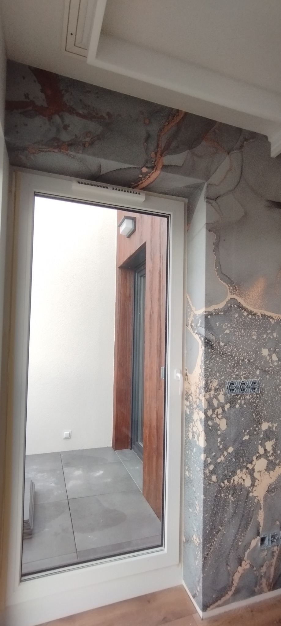 Ściana z dekoracyjną szarą fakturą imitującą marmur, z widocznymi gniazdkami elektrycznymi, biała rama okienna z widokiem na taras z szarą podłogą, drewniana podłoga w pomieszczeniu.