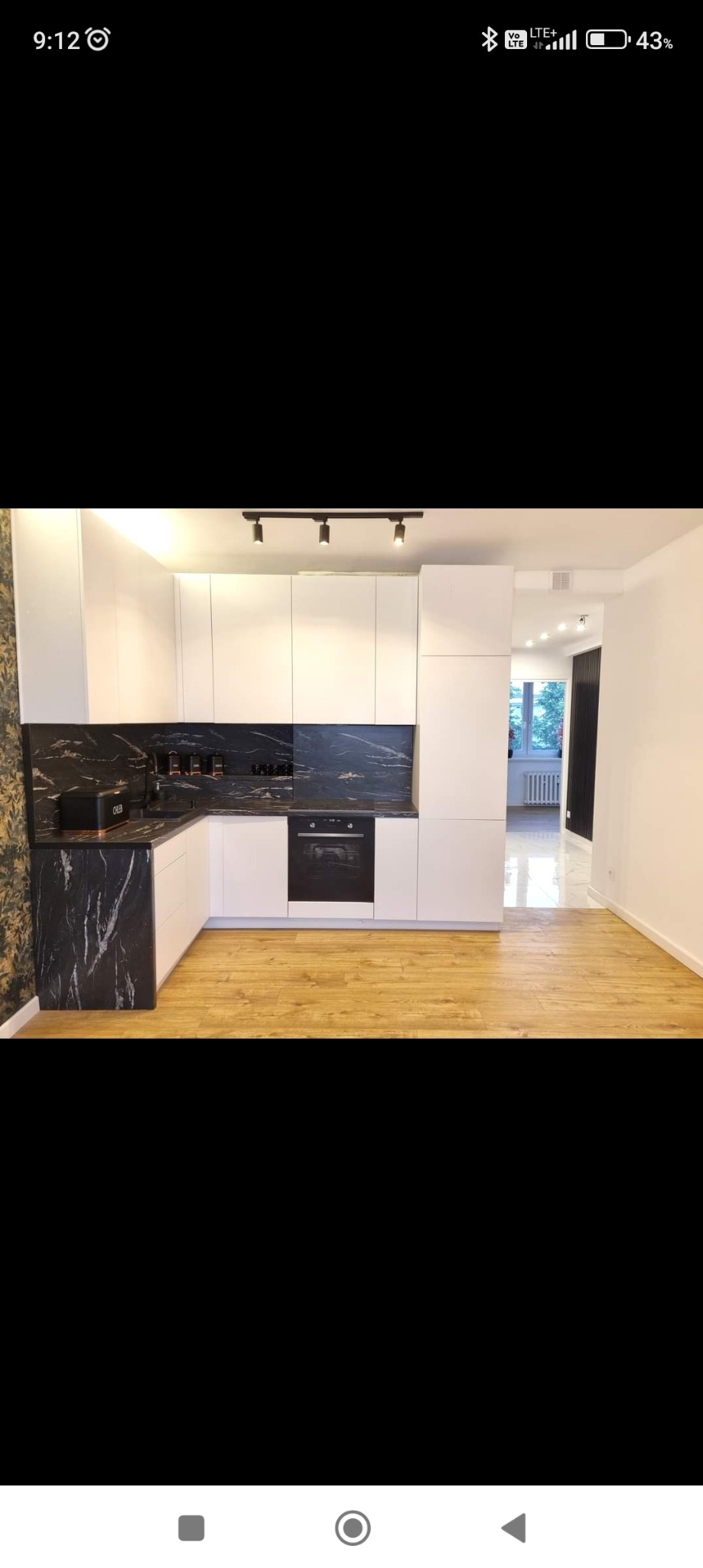 Nowoczesna kuchnia z białymi szafkami i blatem z czarnego marmuru, oświetlenie szynowe, drewniana podłoga, widok na dalszą część mieszkania.