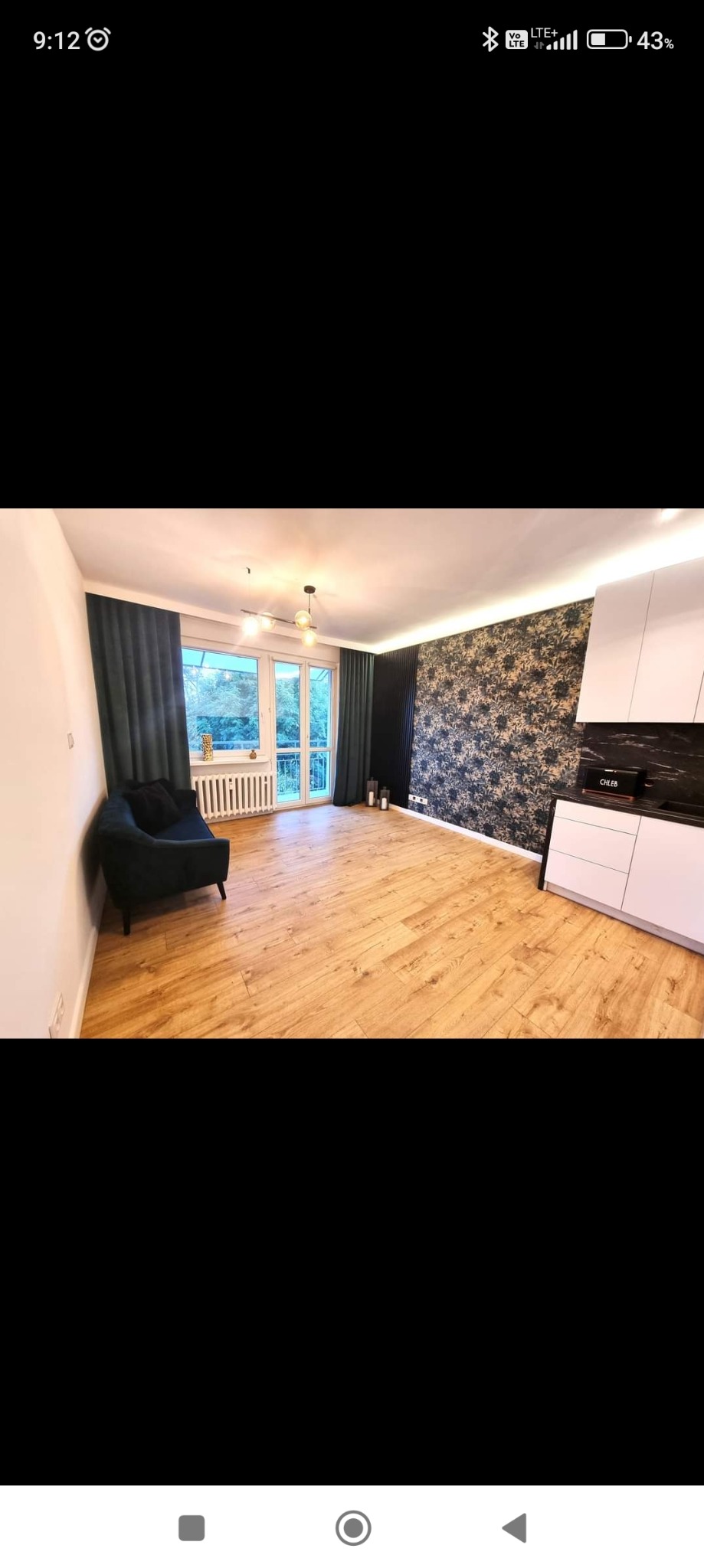 Wnętrze pokoju po remoncie: drewniana podłoga, ściana z tapetą w kwiatowy wzór, ciemnozielone zasłony i sofa, białe meble kuchenne z czarnym blatem.