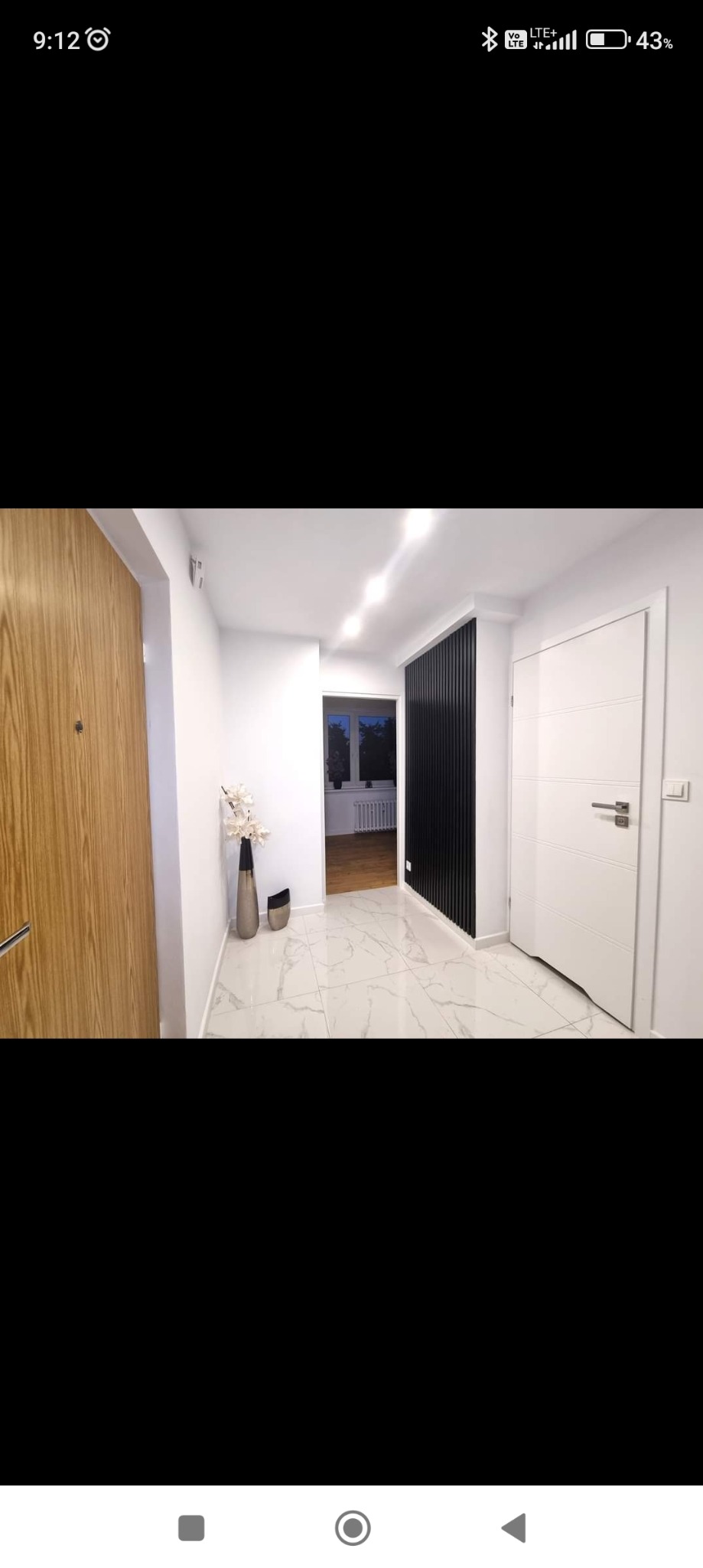 Wykończony korytarz w Opolu z białymi marmurowymi płytkami podłogowymi, drewnianymi drzwiami wejściowymi, białymi ścianami, czarnym panelem dekoracyjnym i białymi drzwiami wewnętrznymi.