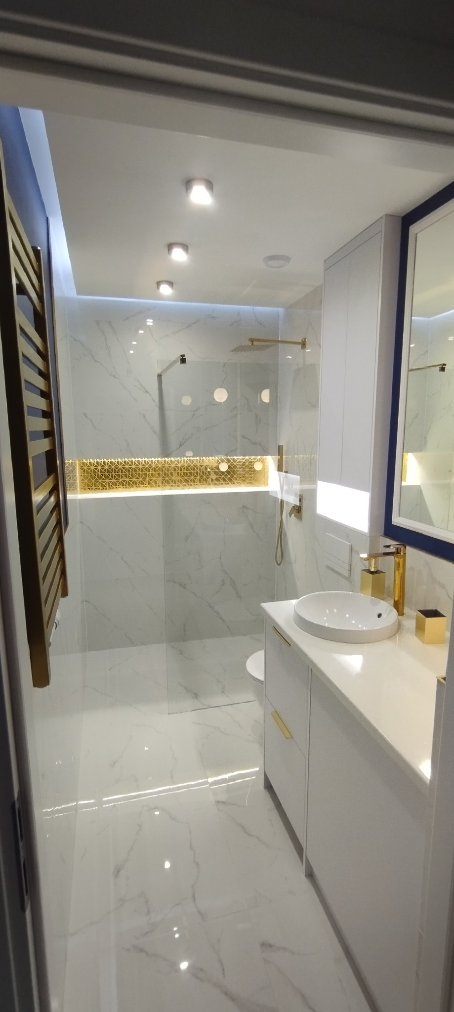 Elegancka łazienka z marmurowymi płytkami, złotymi akcentami i minimalistycznym wyposażeniem, oświetlona ledami.