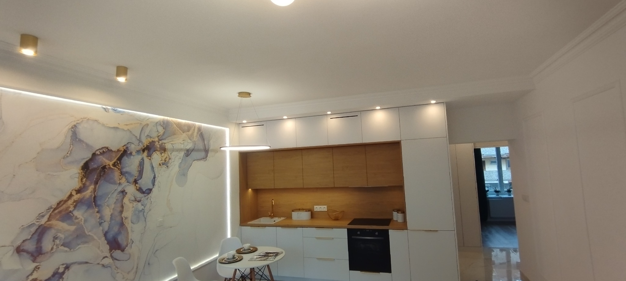 Nowoczesna kuchnia z białymi szafkami, drewnianymi frontami, okrągłym stołem i dekoracyjną tapetą imitującą marmur z niebiesko-złotymi żyłami, oświetlona lampą wiszącą i ledowym paskiem.