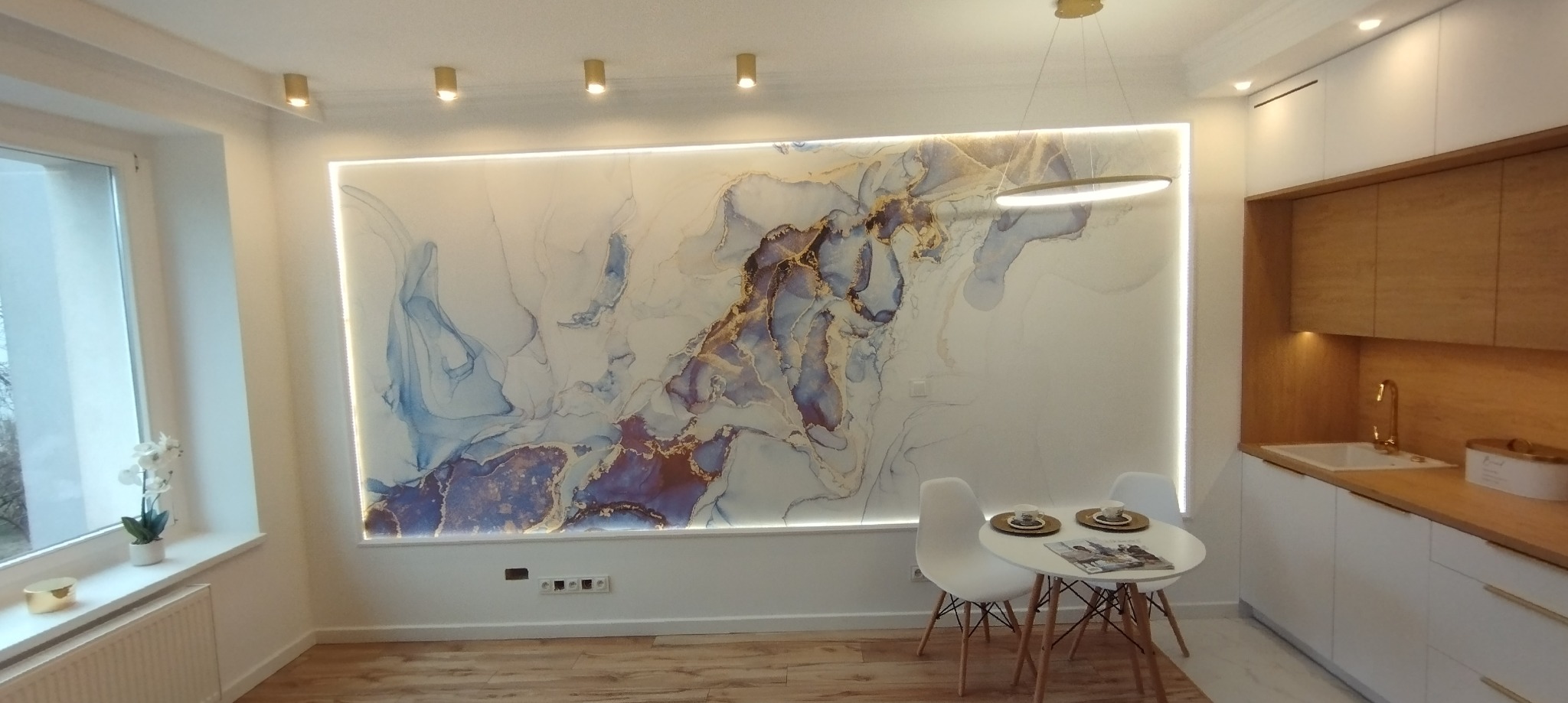 Nowoczesne wnętrze mieszkania z podświetlaną ścianą z abstrakcyjnym wzorem w odcieniach błękitu, złota i fioletu, mały okrągły stolik z dwoma krzesłami, fragment zabudowy kuchennej z drewnianymi...