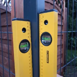 Dwa żółte poziomice przyłożone do słupków ogrodzeniowych, sprawdzające pion, widoczne krople wody na narzędziach.