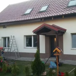 Dom jednorodzinny z nową elewacją w kolorze kremowym, brązowy cokół, czerwona dachówka, pracownik na rusztowaniu maluje ścianę, widoczne elementy ogrodu z roślinami i wózkiem dziecięcym.