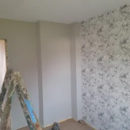 Pokój po częściowym tapetowaniu, jedna ściana w szarym kolorze, druga w tapetę z motywem roślinnym, drabina malarska i narzędzia na podłodze.