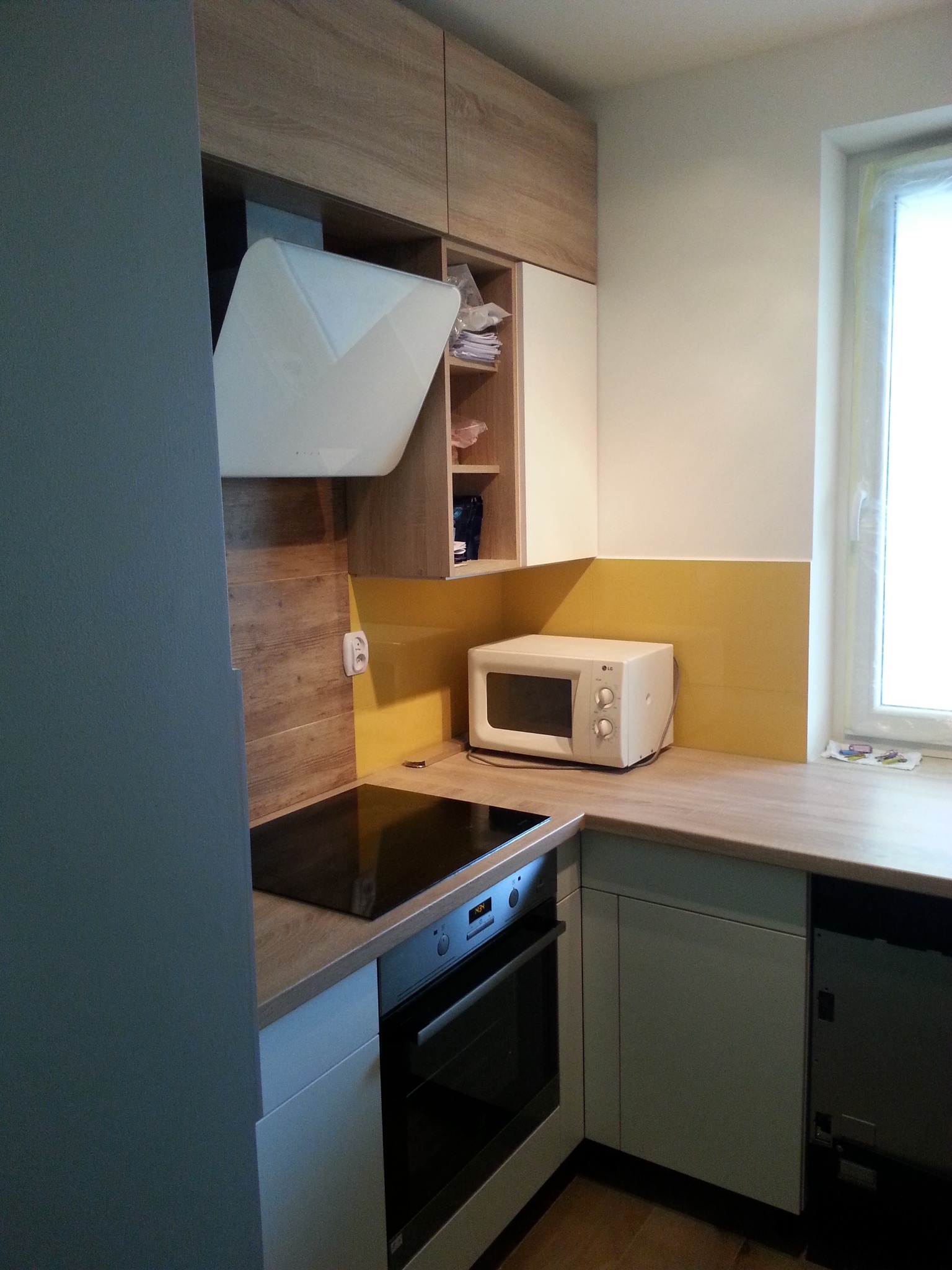 Nowoczesna kuchnia z drewnianymi szafkami górnymi, białymi szafkami dolnymi, żółtym panelem ściennym i czarną płytą grzewczą, widziana z boku.