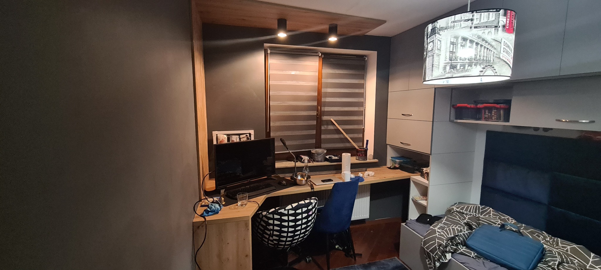 Pokój młodzieżowy z biurkiem z drewnianym blatem, szafkami w zabudowie i lampą z motywem Londynu, widok na okno z roletami.