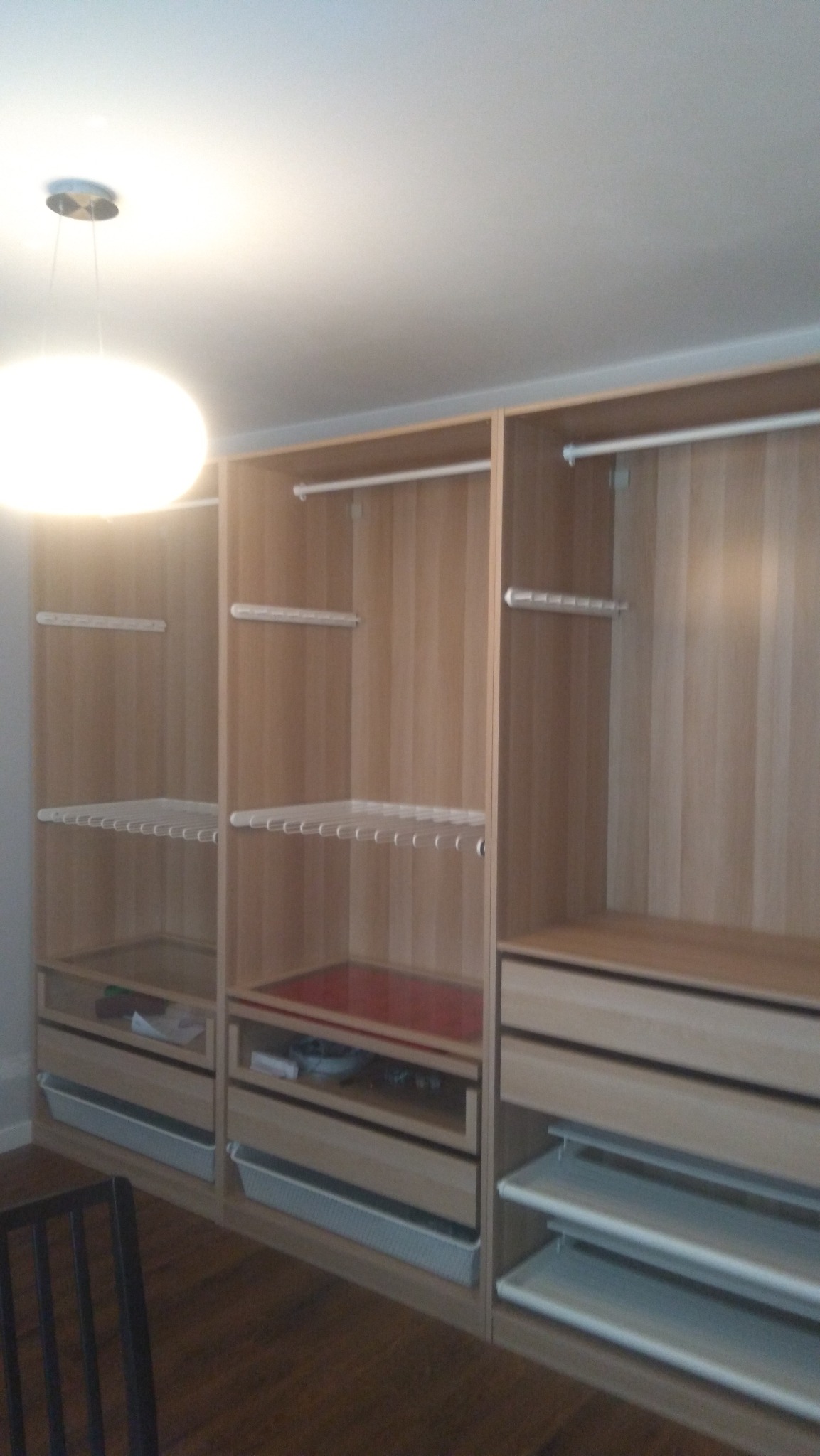 Jasna, drewniana szafa na wymiar z drążkami, półkami drucianymi i szufladami, widoczna część krzesła na pierwszym planie.