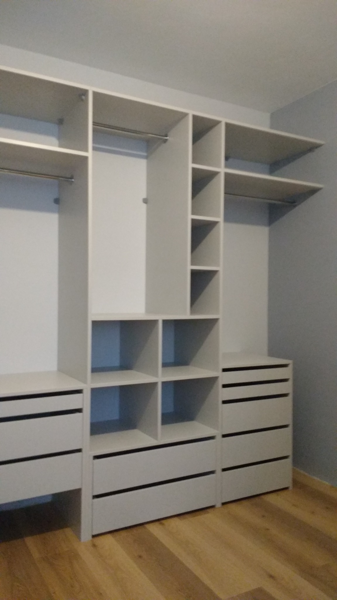 Jasnoszara zabudowa garderoby z szufladami i półkami, widok z przodu na tle parkietu.