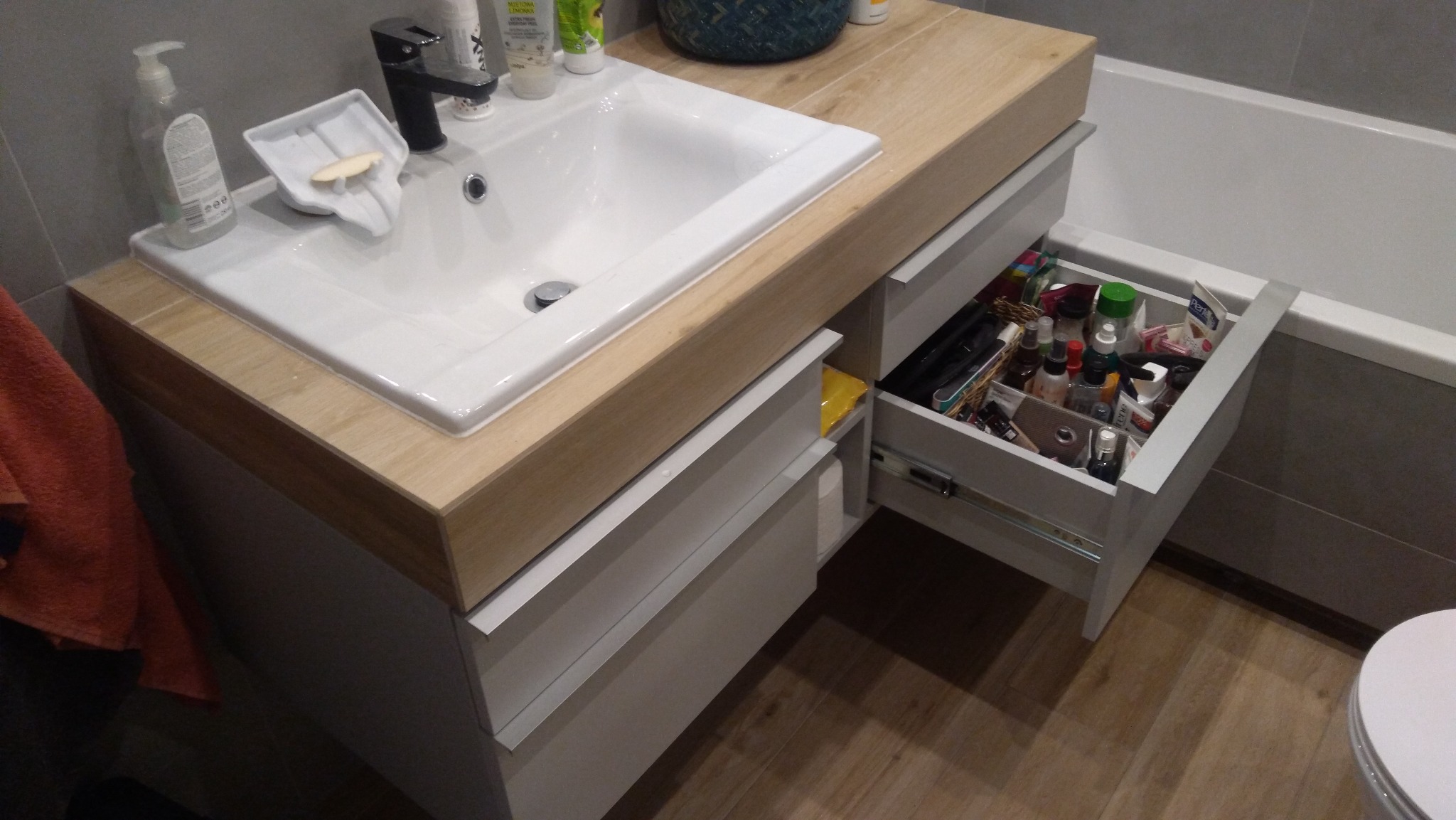 Biała umywalka z czarną baterią, osadzona na szafce z drewnianym blatem, z wysuniętą szufladą wypełnioną kosmetykami i akcesoriami łazienkowymi, widok na fragment wanny i podłogę z paneli.