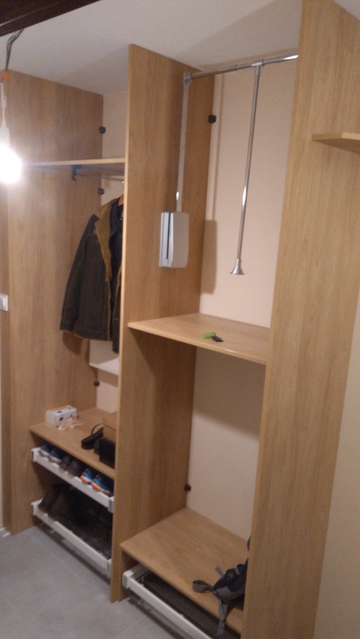 Wnętrze szafy z jasnego drewna z wysuwanymi półkami na buty, wieszakiem na ubrania i metalową rurką do wieszania, widoczne ubrania i akcesoria.