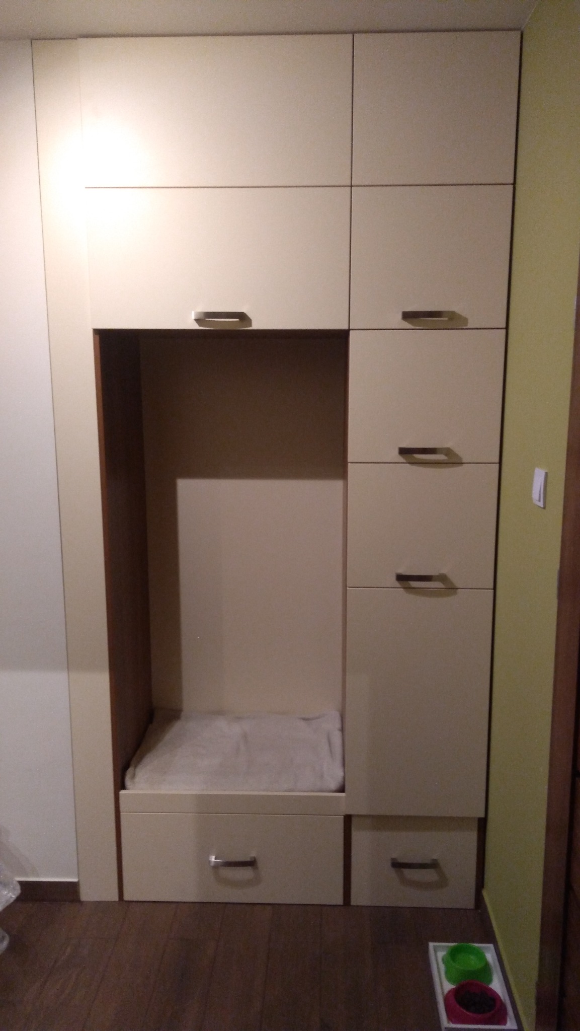 Beżowa szafa z wnęką, w której znajduje się posłanie dla zwierzęcia, widoczne miski z karmą na podłodze.