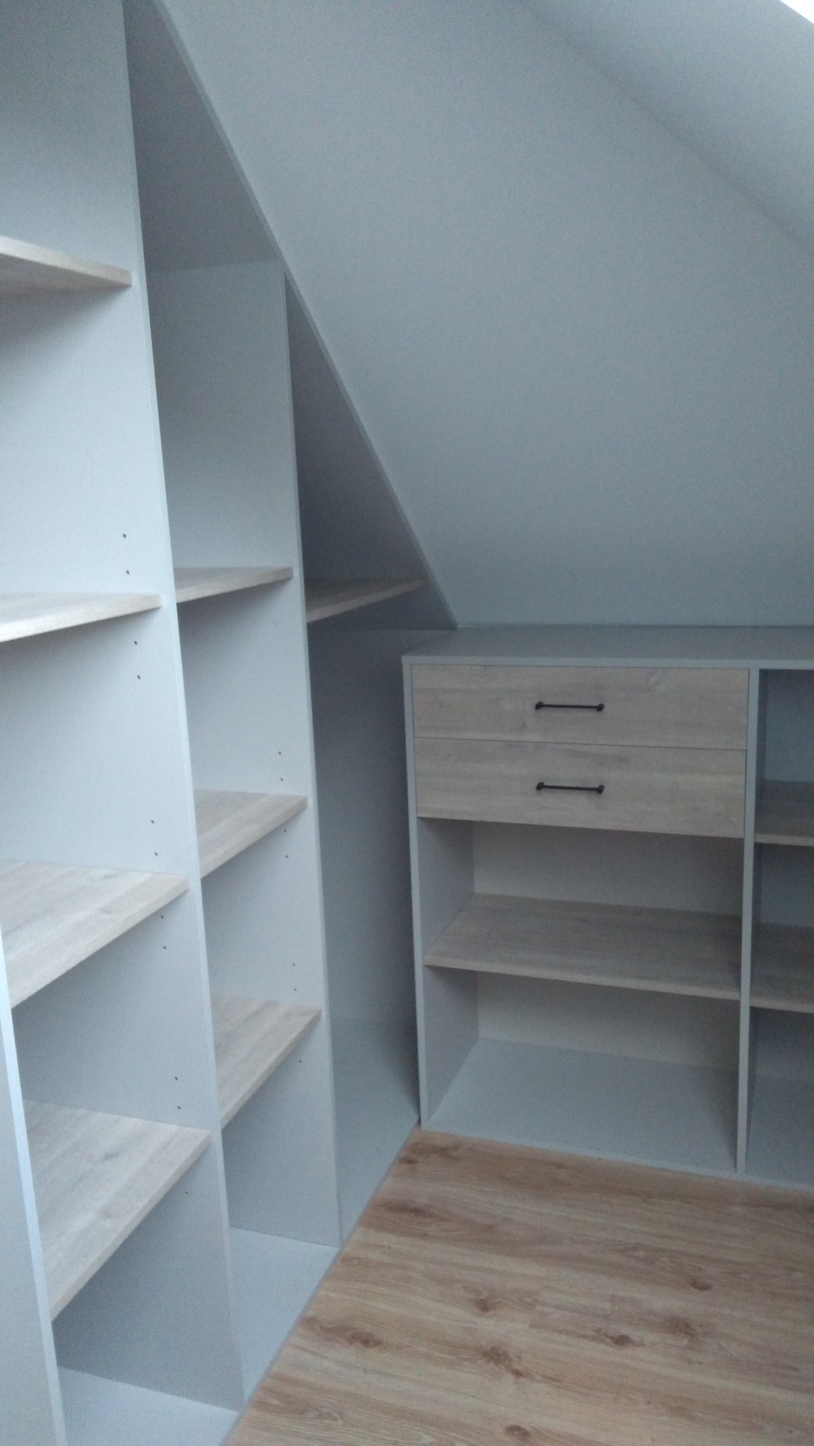 Szara szafa wnękowa z drewnianymi półkami i dwiema szufladami pod skosem dachu, widok z dołu na podłogę z paneli.