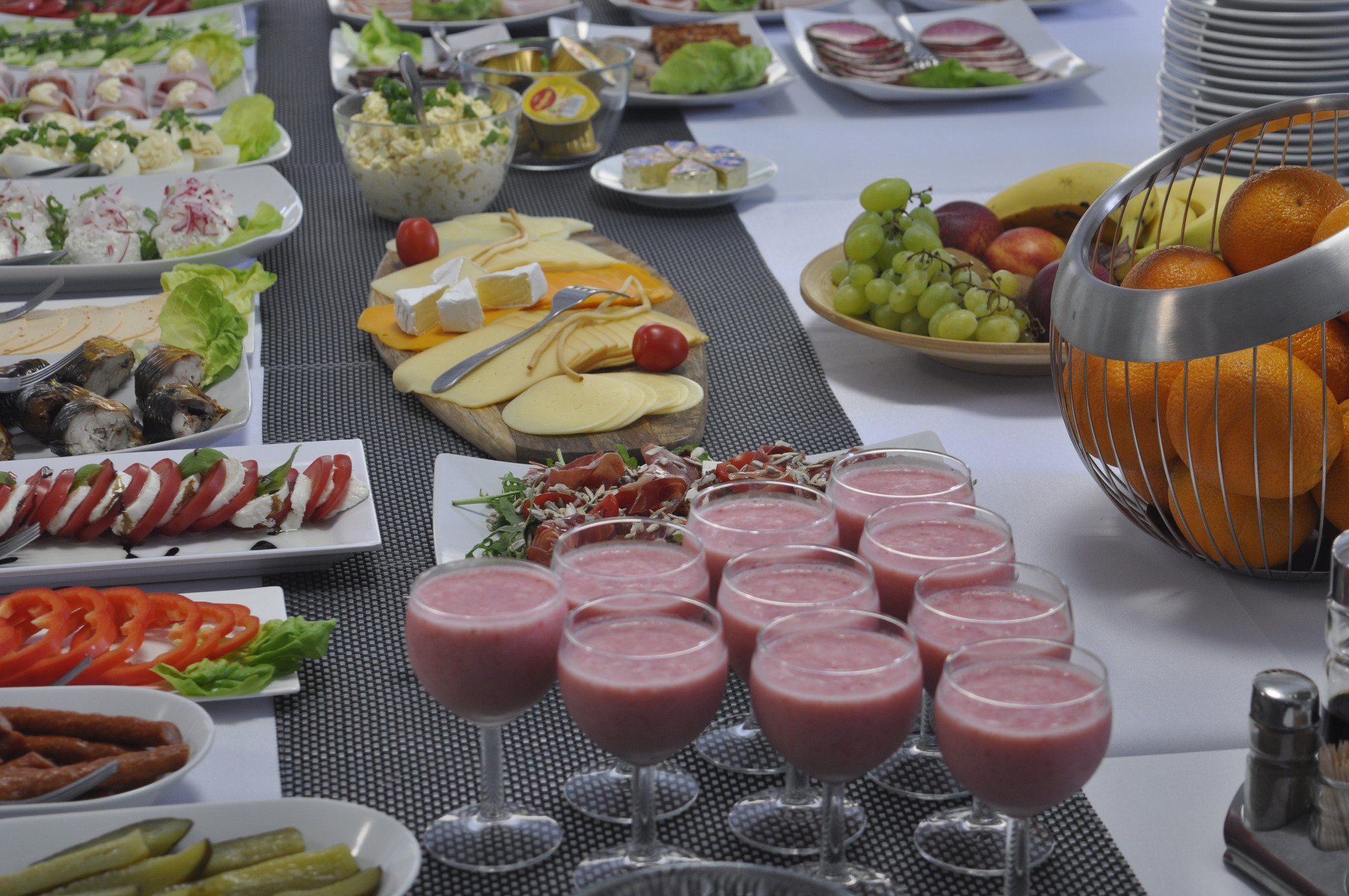 Bogato zastawiony stół z różnorodnymi przekąskami: szklanki z różowym smoothie, deski serów, sałatki, owoce w metalowym koszyku, wędliny i warzywa, tworzące kompozycję cateringu na przyjęciu.