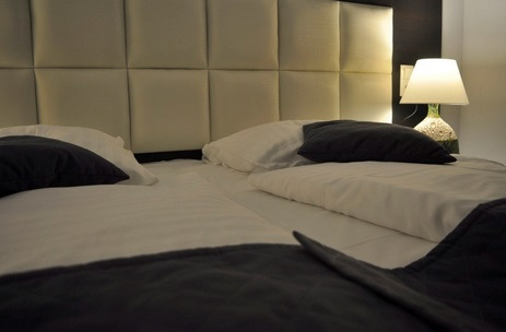 Pokój hotelowy z dwoma łóżkami pojedynczymi, białą pościelą, ciemnymi poduszkami i lampką nocną na stoliku, oświetlony ciepłym światłem.