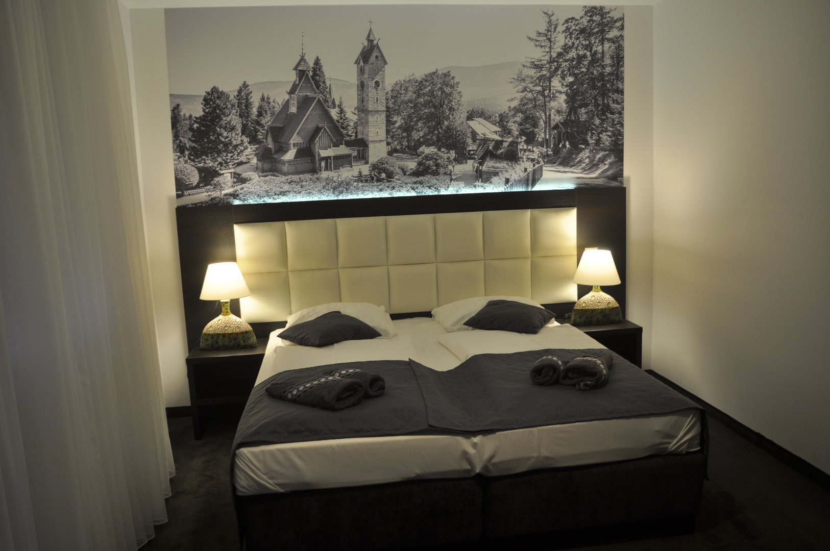 Pokój hotelowy z łóżkiem małżeńskim, tapicerowanym zagłówkiem i czarno-białą fototapetą z motywem górskim i drewnianego kościoła.
