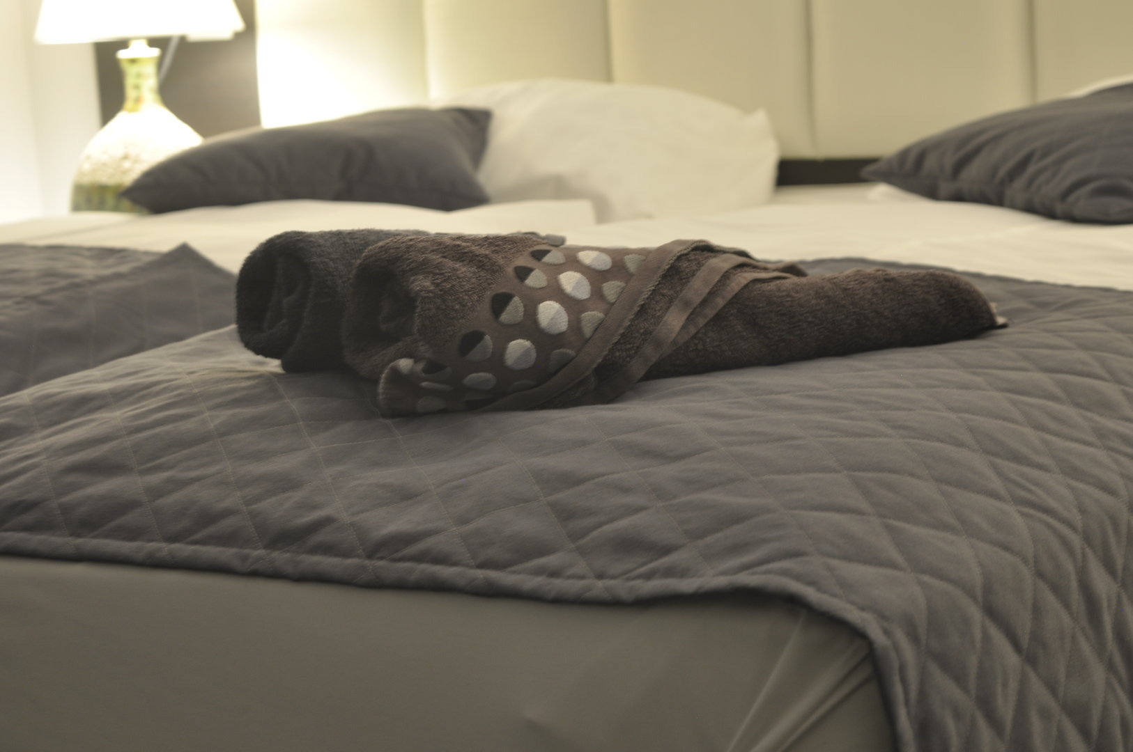 Ułożony szary ręcznik z dekoracyjnymi otworami na szarym łóżku z pikowaną narzutą, w tle lampka nocna.