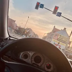 Widok z wnętrza samochodu marki Mercedes na skrzyżowanie z sygnalizacją świetlną i budynkami w tle, widać deskę rozdzielczą z zegarami i kierownicę z logiem marki.