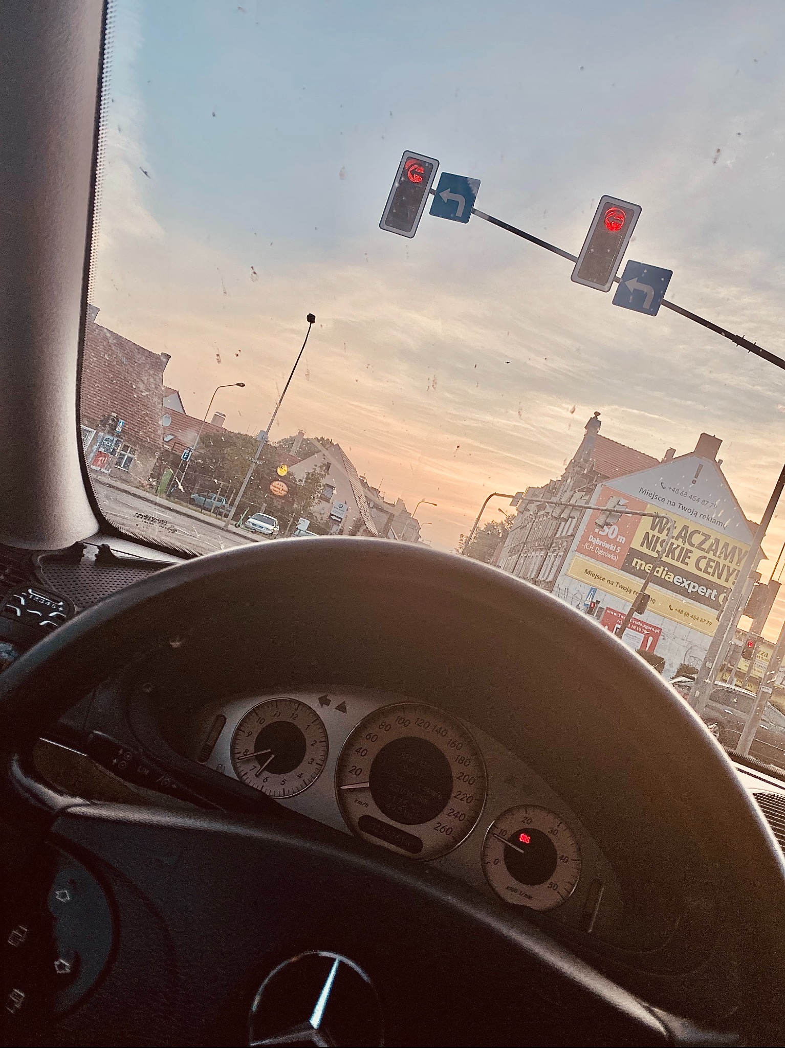 Widok z wnętrza samochodu marki Mercedes na skrzyżowanie z sygnalizacją świetlną i budynkami w tle, widać deskę rozdzielczą z zegarami i kierownicę z logiem marki.