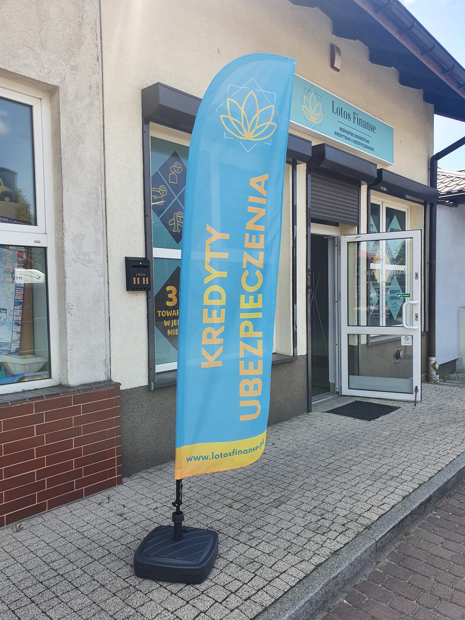 Flaga reklamowa przed biurem Lotos Finanse z napisem Kredyty Ubezpieczenia, widoczny szyld firmy nad wejściem i skrzynka pocztowa.