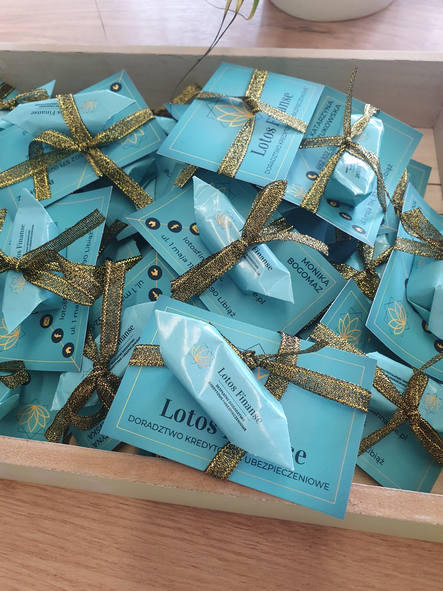 Stos prezentów firmowych w drewnianej skrzynce, zawiniętych w turkusowy papier i przewiązanych złotą wstążką, z nadrukowanym logo firmy 'Lotos Finanse' i danymi kontaktowymi.