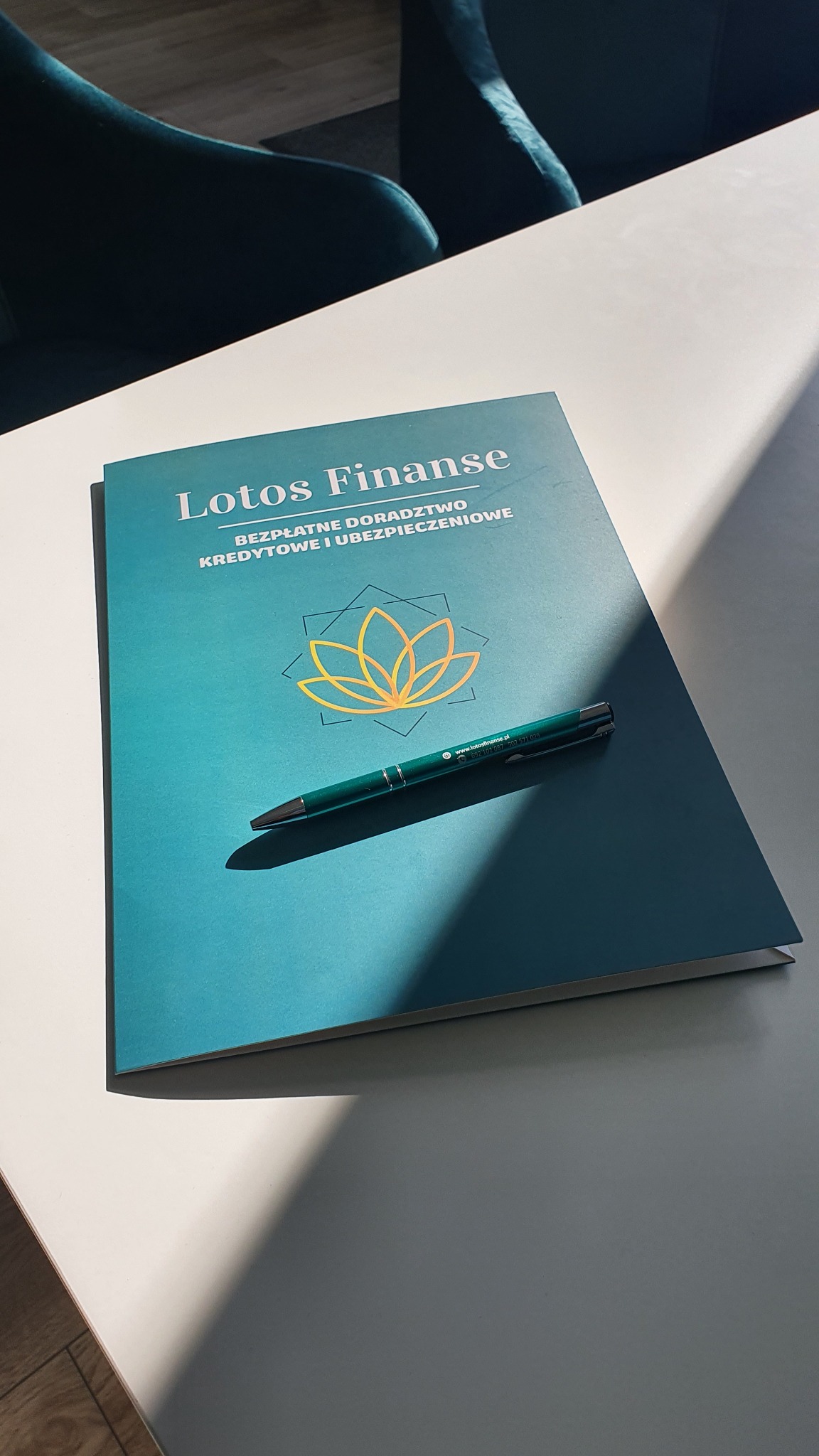 Turkusowy folder z logo Lotos Finanse i napisem 'Bezpłatne doradztwo kredytowe i ubezpieczeniowe' leży na białym stole obok zielonego długopisu.