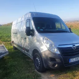 Srebrny Opel Master zaparkowany na trawie obok przyczepy lawety, widok z przodu na tle pola i zabudowań w oddali, słoneczny dzień.