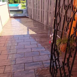 Świeżo ułożona, mokra, brązowa kostka brukowa prowadząca do furtki z ozdobnymi elementami, za którą widać fragment basenu i roślinność ogrodową.