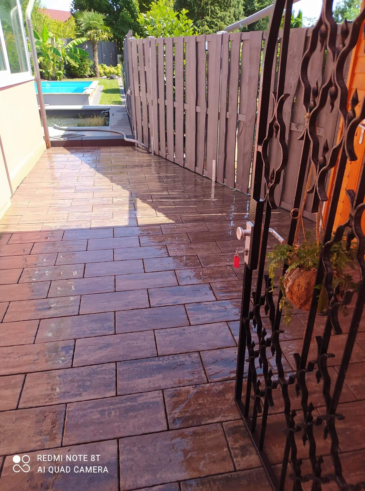 Świeżo ułożona, mokra, brązowa kostka brukowa prowadząca do furtki z ozdobnymi elementami, za którą widać fragment basenu i roślinność ogrodową.