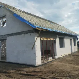 Budowa domu jednorodzinnego w stanie surowym otwartym, widoczne ściany z bloczków, drewniana konstrukcja dachu z membraną, stalowe podpory szalunkowe.