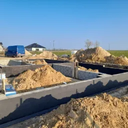 Wykopany fundament z izolacją pionową, częściowo zasypany ziemią, widoczne bloczki betonowe, styropian do izolacji termicznej i zaparkowany samochód dostawczy w tle.