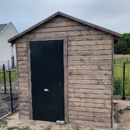 Mała drewniana budowla gospodarcza z czarnymi drzwiami, widoczna na tle ogrodzenia i domu.