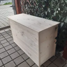 Surowa, jasna drewniana skrzynia ogrodowa na podwórku z kostki brukowej, z zielonym bluszczem w tle.