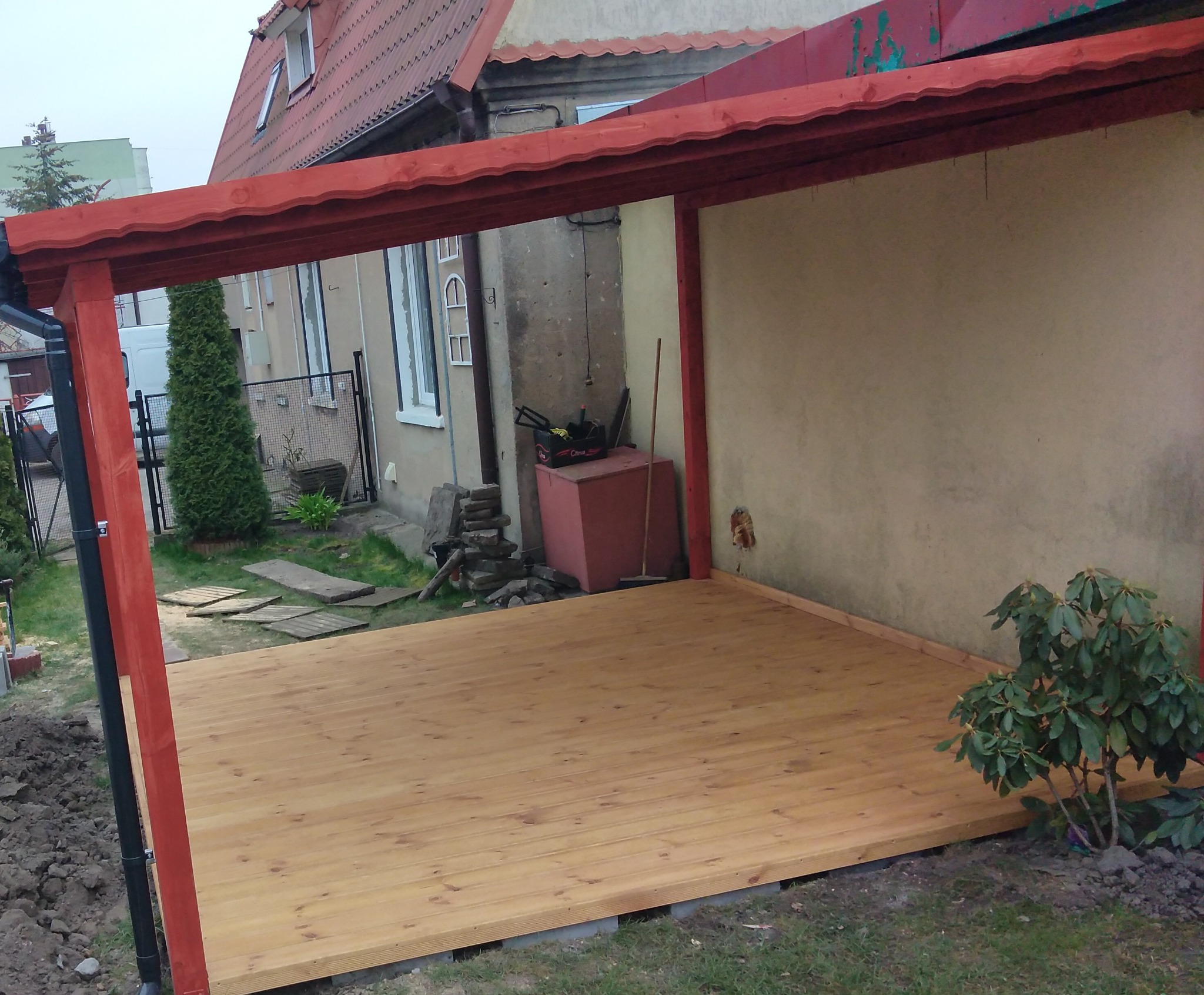 Zadaszona, świeżo wybudowana konstrukcja z drewna pomalowanego na czerwono, z widoczną nową podłogą z desek, narzędzia majsterkowicza i roślinność w tle.