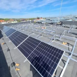 Montaż paneli fotowoltaicznych na dachu budynku, widoczne rzędy paneli i aluminiowa konstrukcja wsporcza, w tle zabudowa miejska i błękitne niebo.