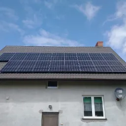 Panele słoneczne na brązowym dachu dwuspadowym domu jednorodzinnego z szarą elewacją, widoczne okno z zielonymi zasłonami, antena satelitarna i fragment błękitnego nieba z chmurami.