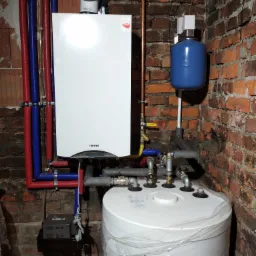 Biały piec CO marki Termet zamontowany na tle ceglanej ściany, widoczne rury w izolacji, zbiornik wyrównawczy i zasobnik ciepłej wody z folią ochronną.