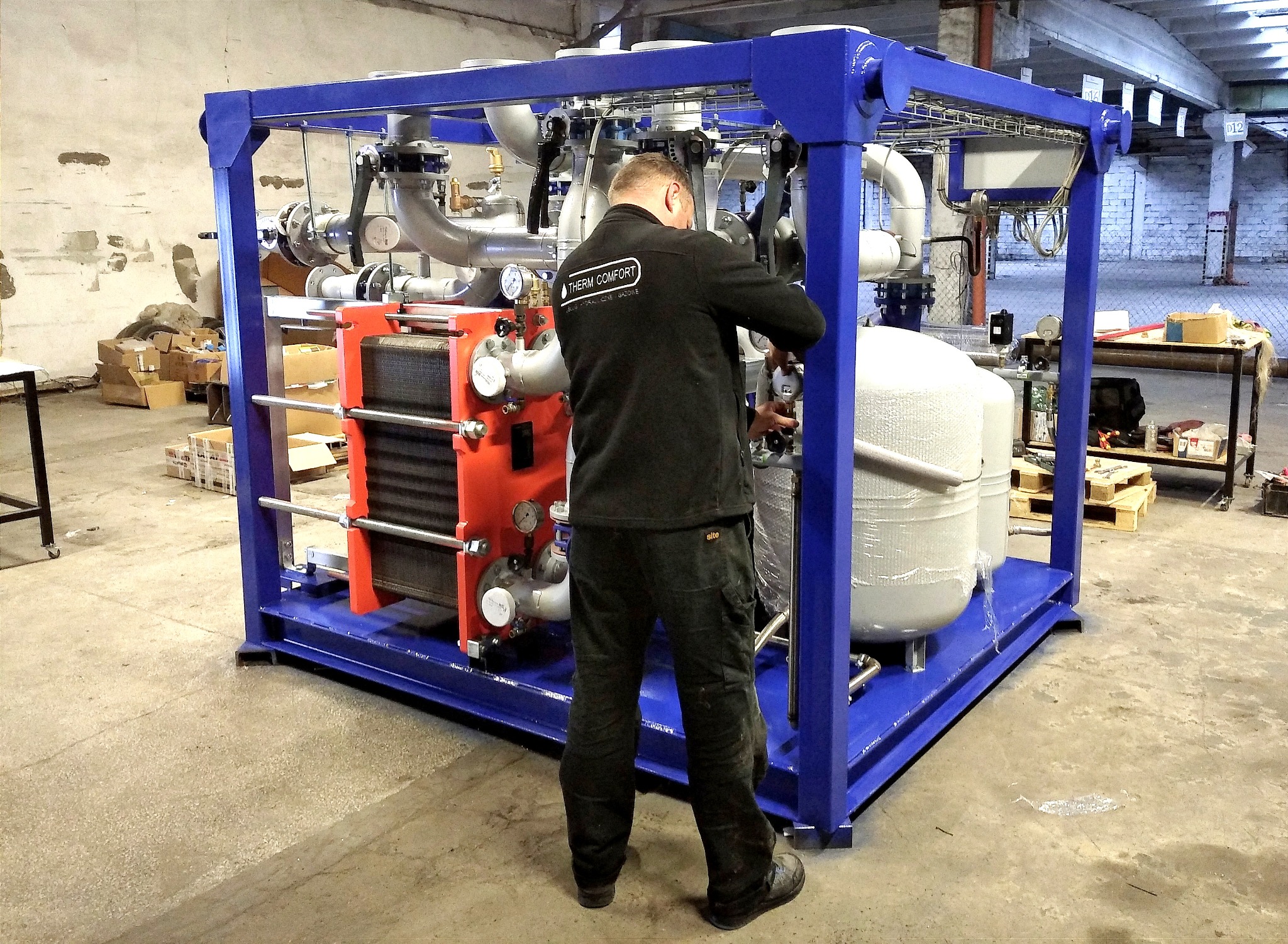 Mężczyzna w czarnej bluzie z logo firmy instalacyjnej, reguluje zawór w przemysłowym wymienniku ciepła z orurowaniem, zamontowanym na niebieskiej ramie w hali.