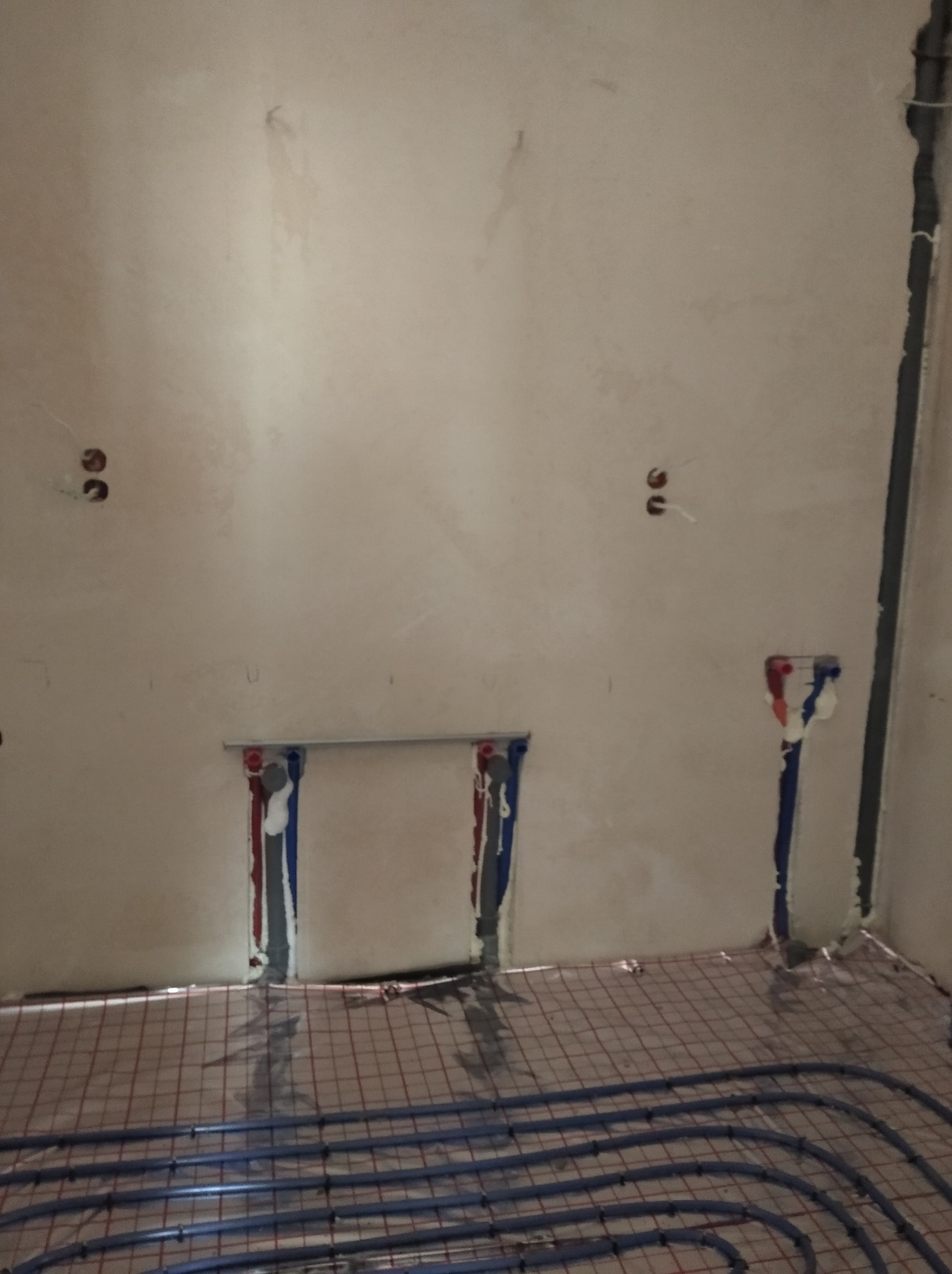Surowa ściana z widocznymi otworami na instalacje wodne i elektryczne, na podłodze ułożona siatka grzewcza z niebieskimi rurkami ogrzewania podłogowego.