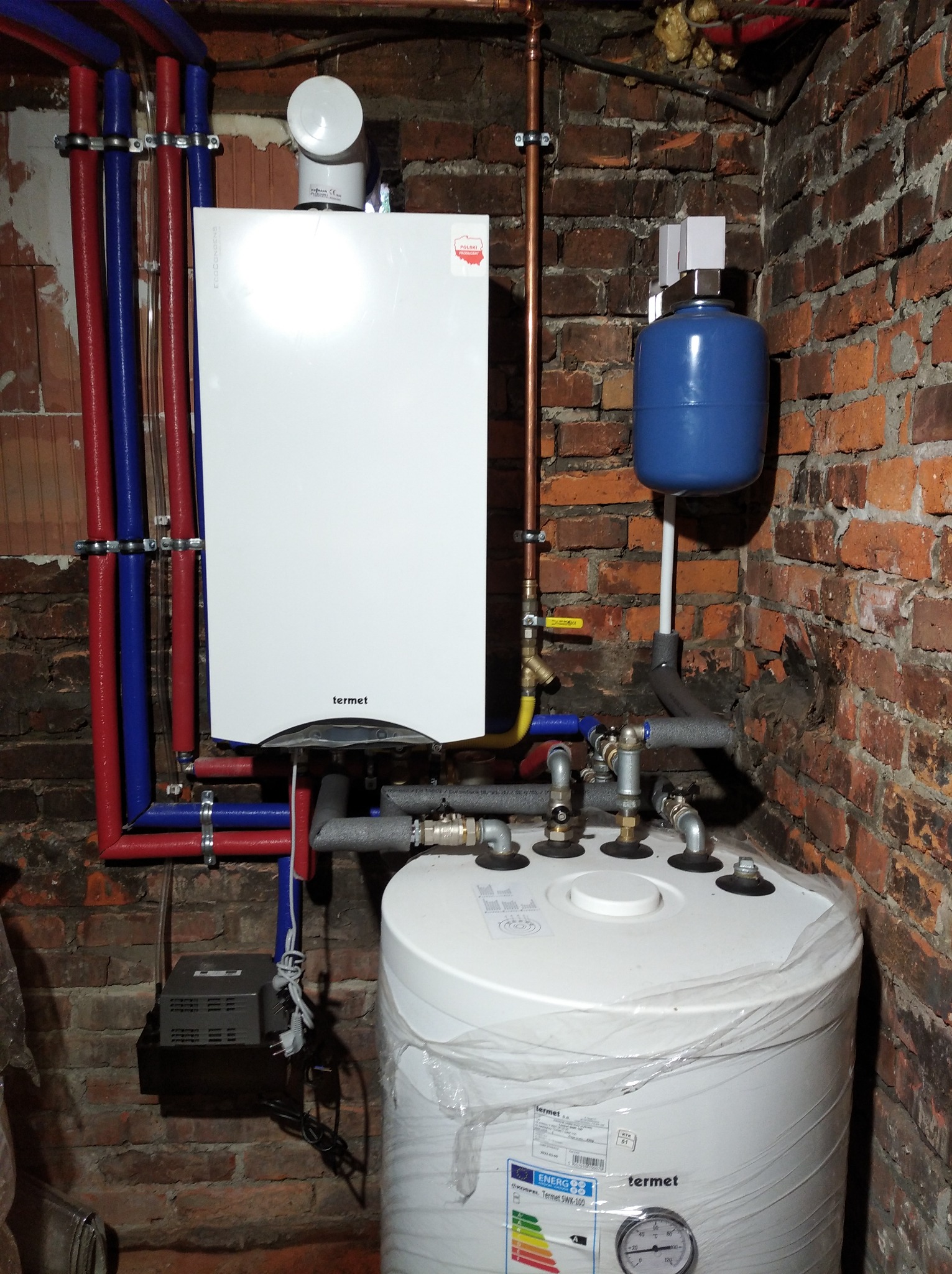Biały piec CO marki Termet zamontowany na tle ceglanej ściany, widoczne rury w izolacji, zbiornik wyrównawczy i zasobnik ciepłej wody z folią ochronną.