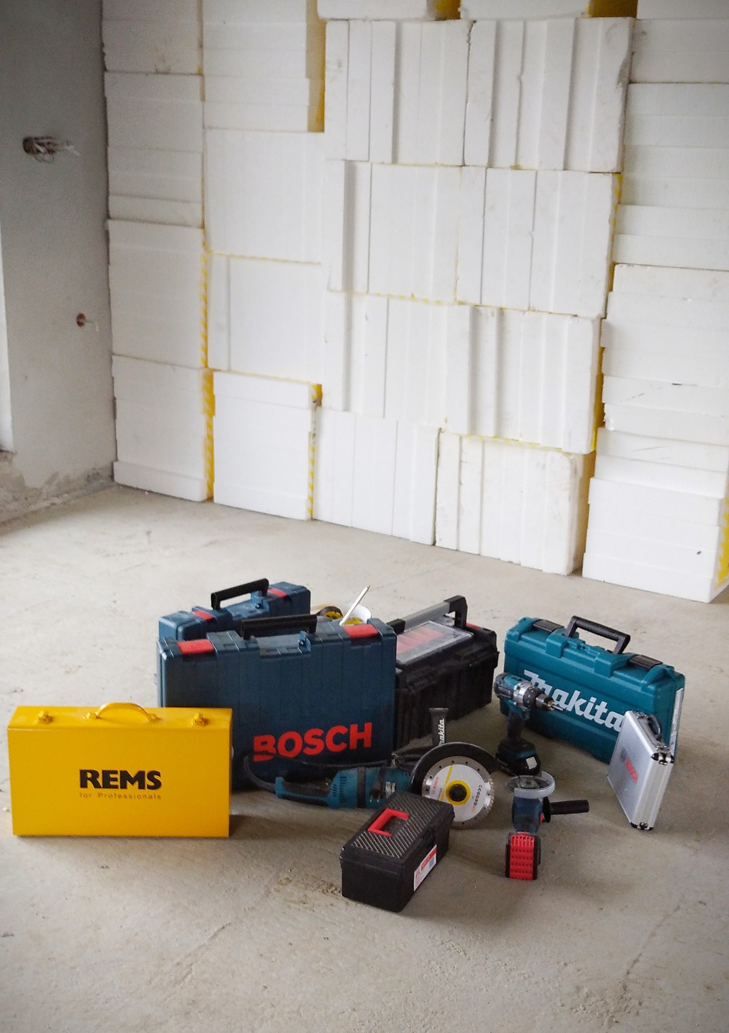 Stos białych płyt izolacyjnych w surowym pomieszczeniu, narzędzia i skrzynki narzędziowe marek REMS, Bosch i Makita na podłodze.
