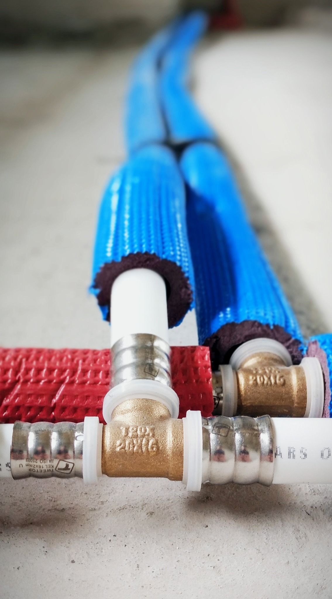 Połączenie rur instalacji wodnej i grzewczej: białe rury z mosiężnymi złączkami, niebieskie i czerwone otuliny izolacyjne na szarym betonowym podłożu w trakcie montażu.